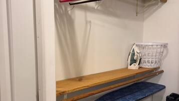A compact room with a wooden shelf, a blue patterned bench, a hanging laundry rack, and a white laundry basket.