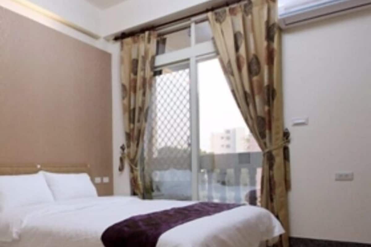 A bedroom with a bed, a window with curtains, and a view of buildings outside.