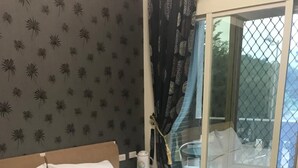 Un lit bien fait, avec des draps blancs et un plaid violet foncé, une table de chevet avec une lampe, et une fenêtre donnant sur l’extérieur.