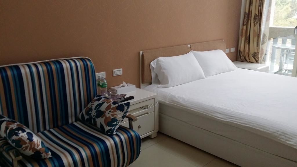 Habitación de hotel con un sofá a rayas, una cama con ropa blanca, una mesita de noche con una botella y una caja de pañuelos, y una ventana con cortinas.