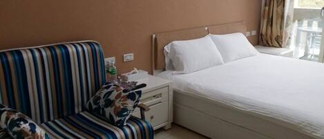 Habitación de hotel con un sofá a rayas, una cama con ropa blanca, una mesita de noche con una botella y una caja de pañuelos, y una ventana con cortinas.