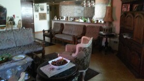 Un salon avec un canapé, des fauteuils et une table basse. Au fond, on aperçoit un coin cuisine avec un réfrigérateur et un évier.