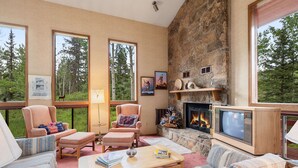 Un salon chaleureux avec une cheminée en pierre, des fauteuils confortables et de grandes fenêtres donnant sur une forêt.