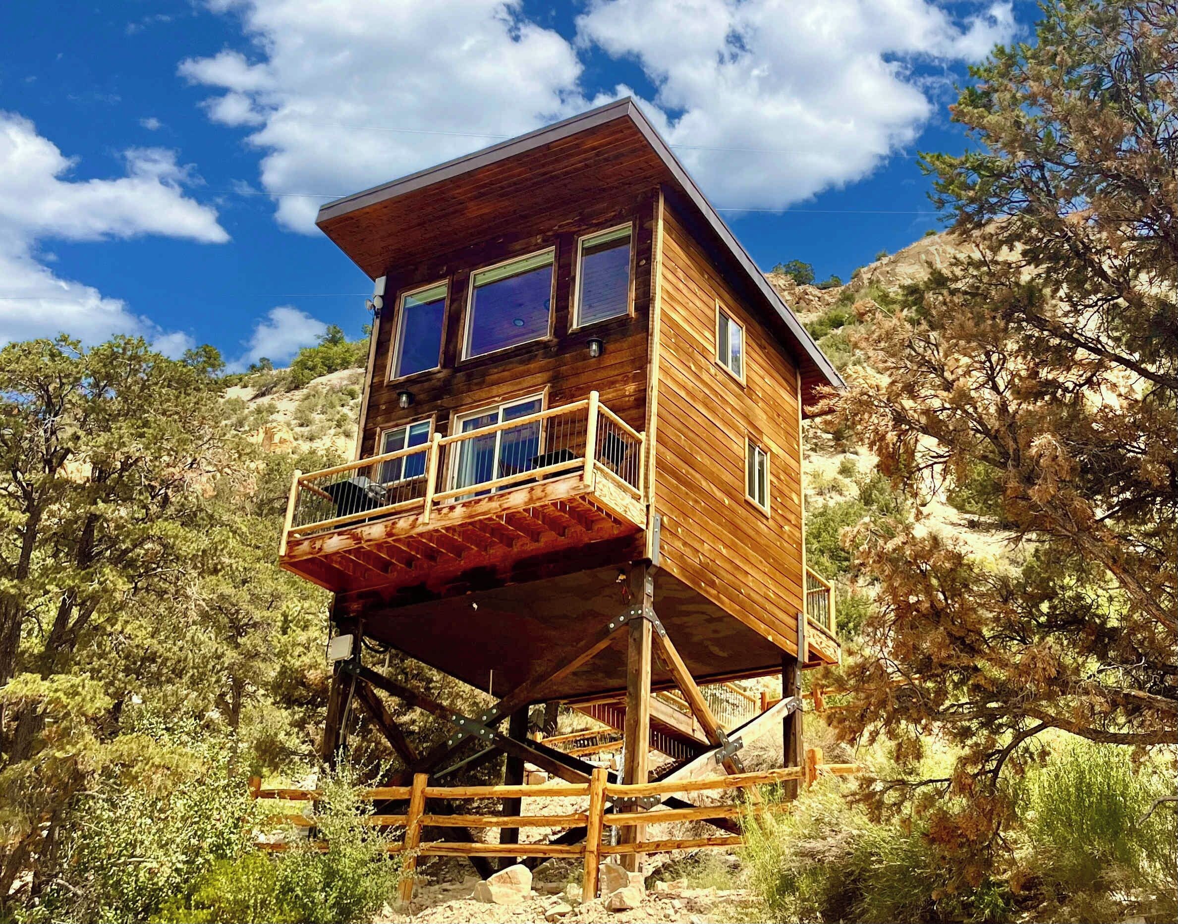 A wooden cabin with a balcony, situated on stilts amidst a rocky landscape with trees.