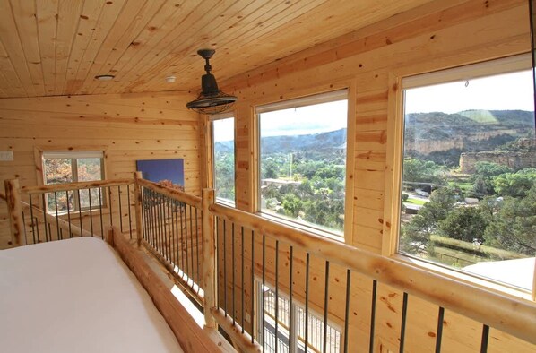 A wooden cabin room with a bed, a window, and a view of a mountainous landscape.