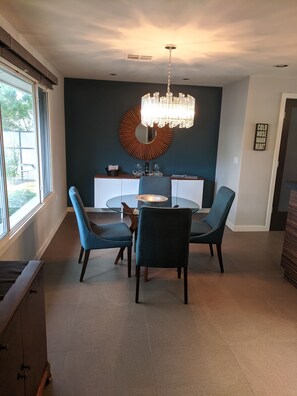 A modern dining area with a round glass table, four chairs, a chandelier, and a decorative wall piece.