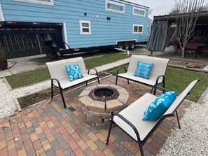 An outdoor patio with two chairs and a fire pit, surrounded by a brick patio and a blue house in the background.