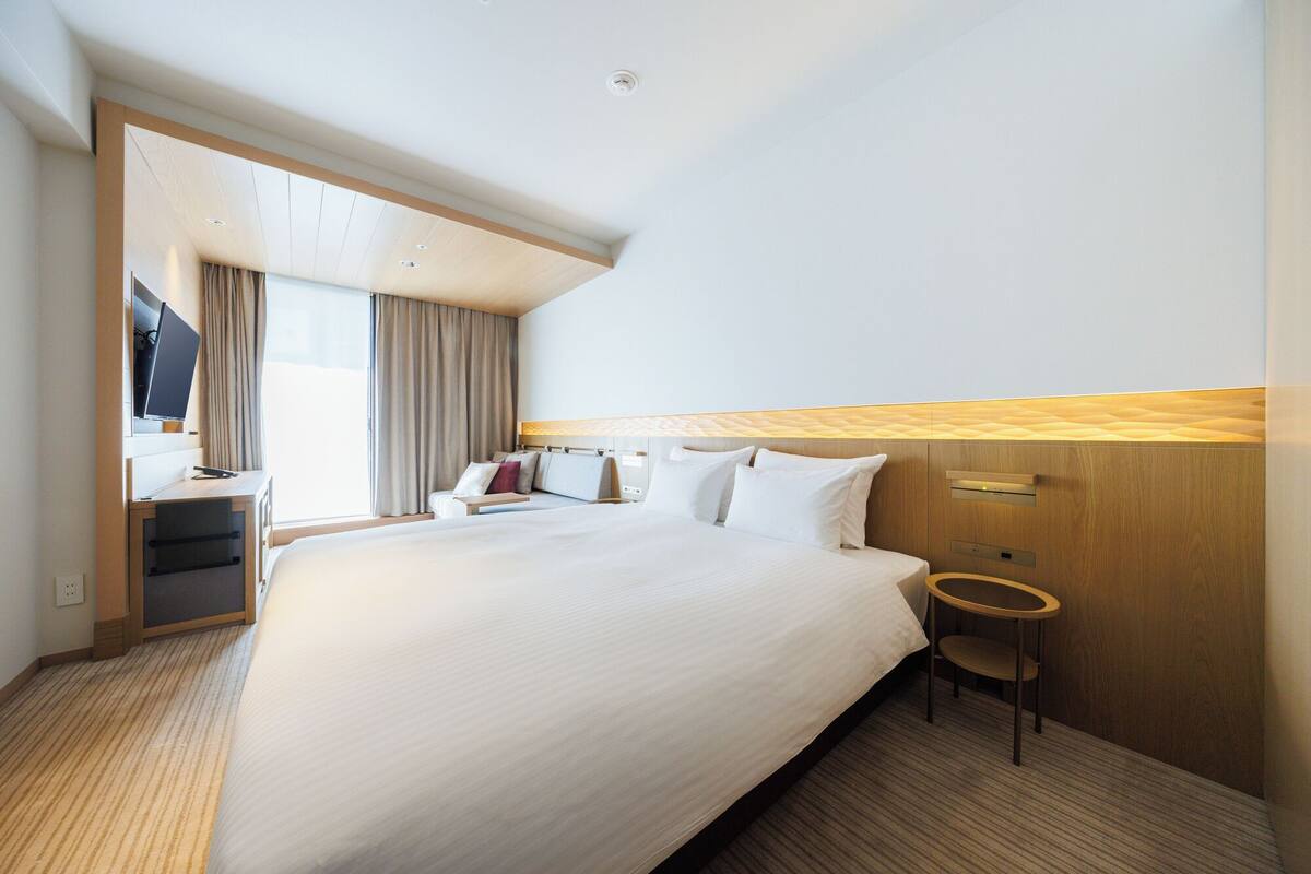 Quarto de hotel com uma cama grande, uma escrivaninha com televisão de tela plana, uma cadeira e uma mesinha.