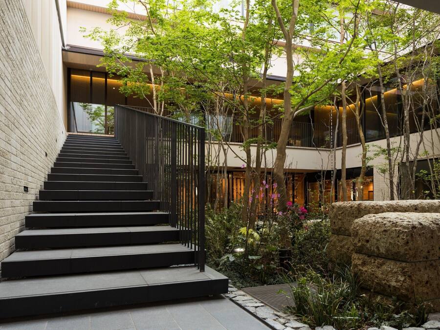 A modern building with a staircase leading to an upper level, surrounded by greenery and stone features.