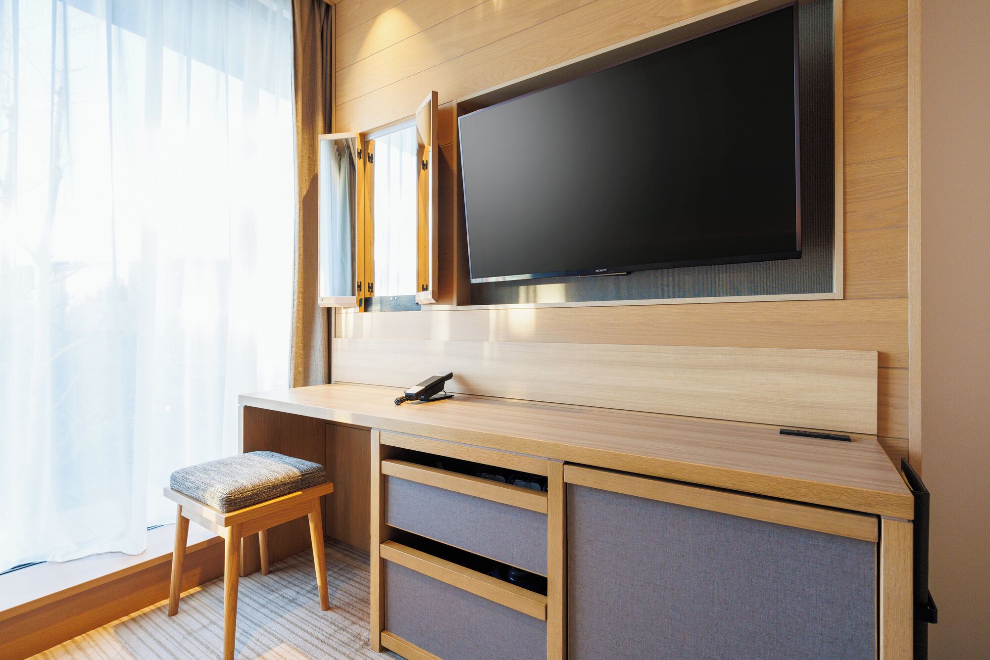 Una habitación de hotel moderna con un escritorio de madera, un televisor de pantalla plana, una silla y un ventanal con cortinas translúcidas.