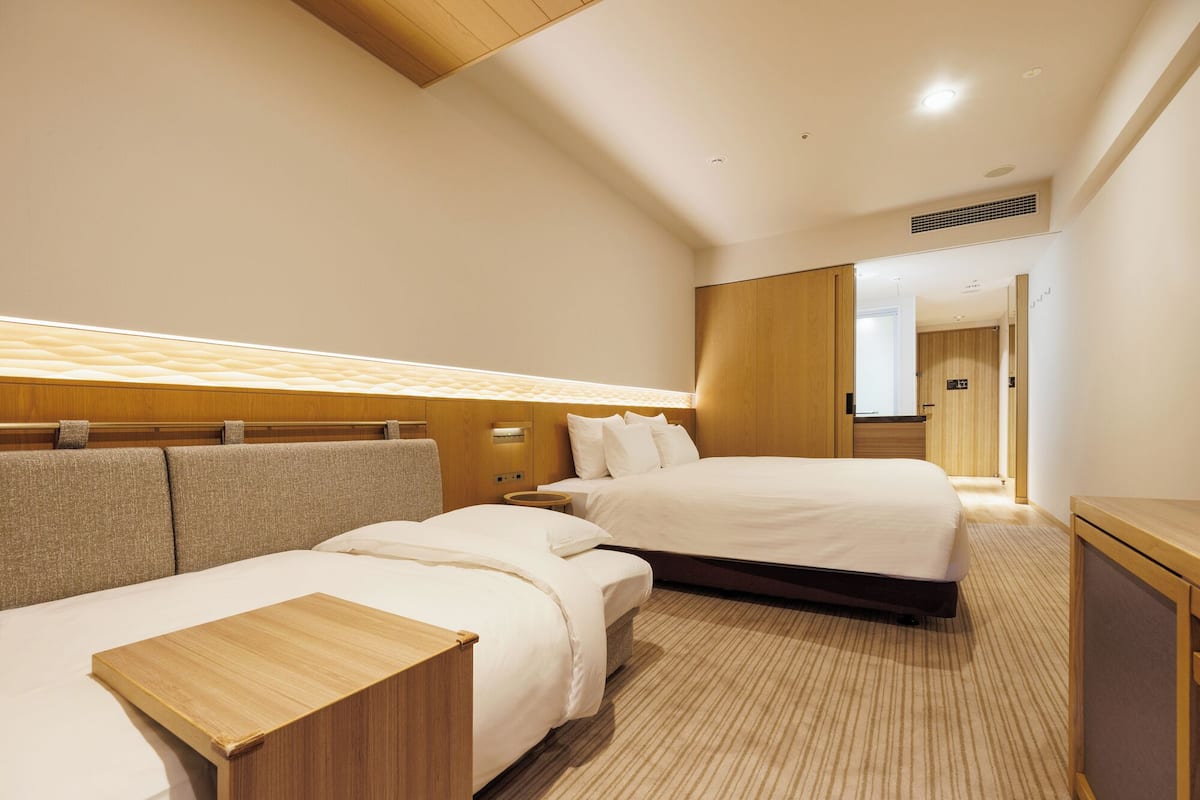 Quarto de hotel com duas camas, cabeceira de madeira, mesa de cabeceira e guarda-roupa.