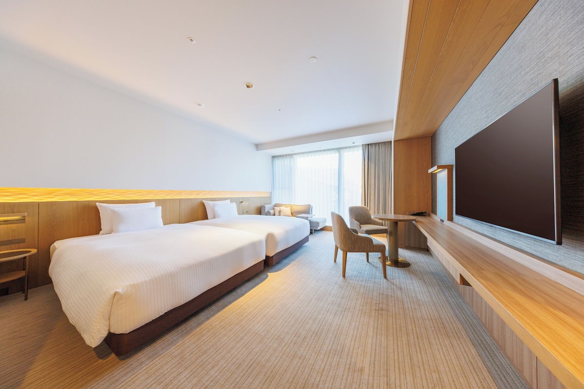 Quarto de hotel com uma cama grande, uma mesa, uma cadeira, uma televisão e uma janela com cortinas.