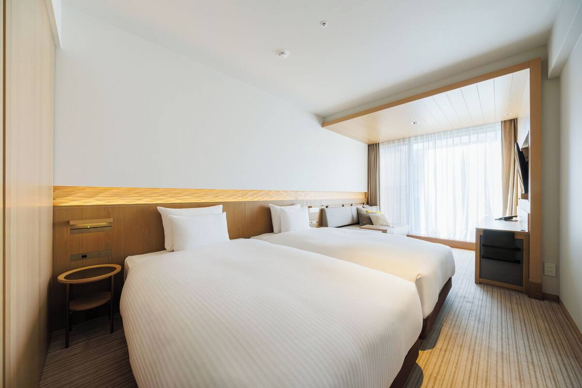 Quarto de hotel com duas camas, uma escrivaninha, uma cadeira e uma janela grande.