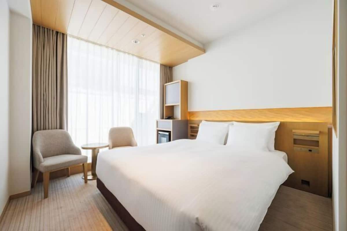 Quarto de hotel com uma cama grande, uma cadeira, uma mesa pequena, uma televisão e uma escrivaninha com abajur.