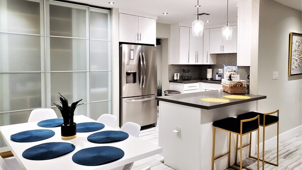 Een moderne keuken met een centraal eiland, witte kasten en een eethoek met een witte tafel en blauwe kussens.