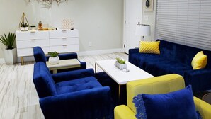 Een moderne woonkamer met een witte bank, blauwe fauteuils, een gele fauteuil en een blauwe bank met gele kussens. Er staat een witte salontafel, een witte dressoir met decoratieve objecten, en een plant.