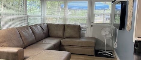 A room with a sectional sofa, a small chair, a standing fan, and a window with blinds.
