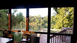 Ein Zimmer mit großem Fenster mit Blick auf See und Wald. Vor der Tür befindet sich eine Holzterrasse mit Geländer und ein Stuhl im Zimmer.