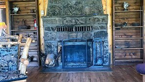 Interior de uma cabana de madeira com lareira de pedra, prateleiras de madeira e uma pintura emoldurada de uma paisagem.