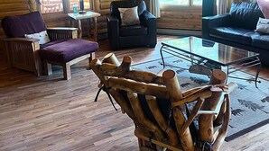 Interior de uma cabana de tronco com piso de madeira, um sofá de couro preto, uma cadeira de madeira e uma mesa de centro de vidro.
