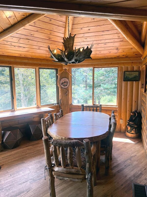 Interior de una cabaña de madera con una mesa redonda y sillas, ventanas amplias y una lámpara en el techo.