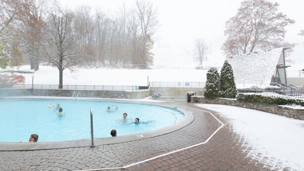 Une piscine où l’on voit des personnes, entourée d’un chemin pavé en briques et d’un sol recouvert de neige.