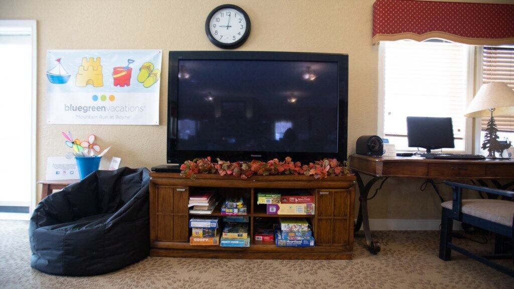 Una sala de estar con una televisión de pantalla plana, un sillón de saco, una estantería con juegos de mesa, un reloj y una ventana con persianas.