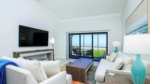 A modern hotel room with a flat-screen TV, a sofa, a coffee table, and a balcony with a view of the ocean.