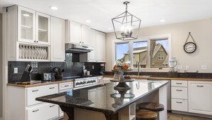 Una cocina con gabinetes blancos, una isla grande con taburetes, una lámpara colgante y una ventana con vista a una casa.