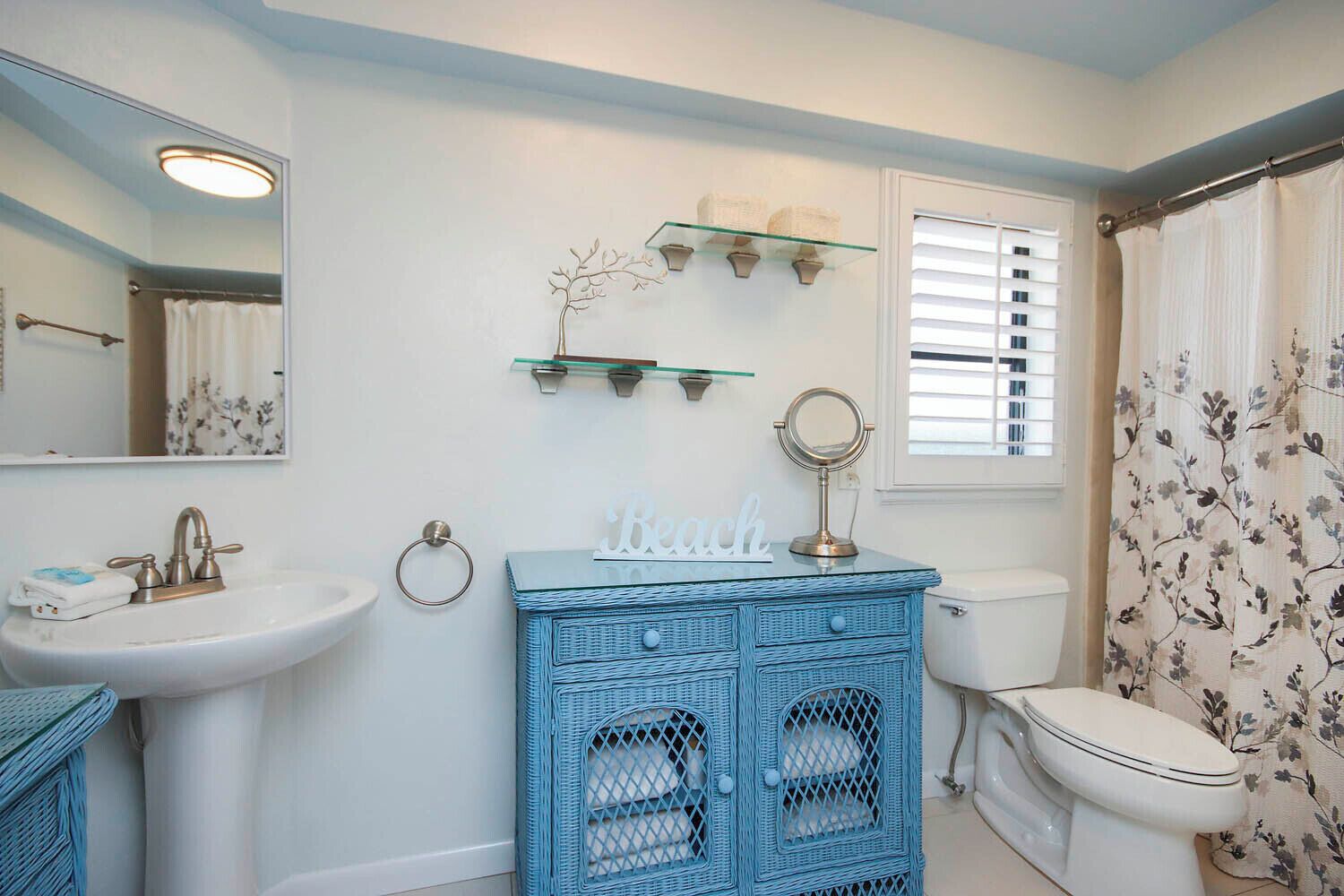 Un baño con lavamanos blanco, tocador de mimbre azul, inodoro, espejo y ventana con persianas.