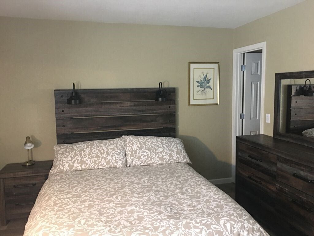 A bedroom with a wooden headboard, a bed with patterned bedding, a nightstand with a lamp, a dresser, and a framed picture on the wall.