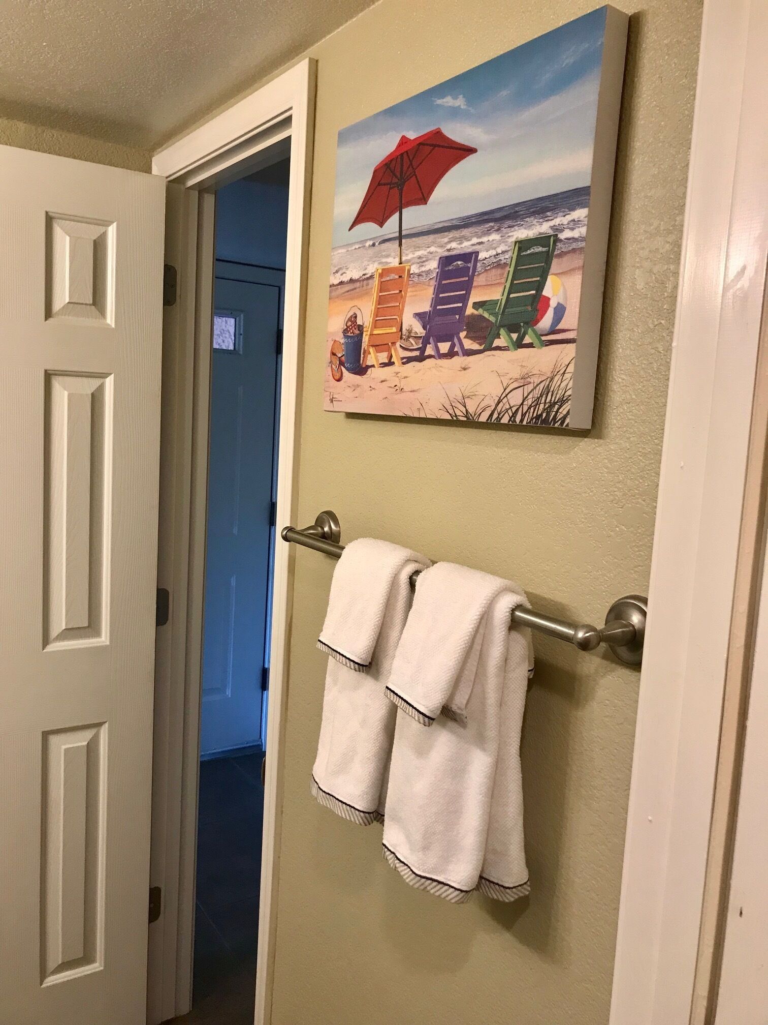 A bathroom with a towel rack holding two white towels, a painting of beach chairs and an umbrella, and a door leading to another room.