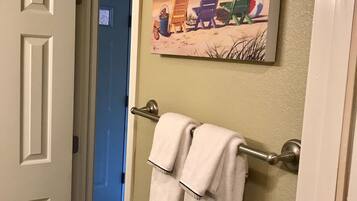 A bathroom with a towel rack holding two white towels, a painting of beach chairs and an umbrella, and a door leading to another room.