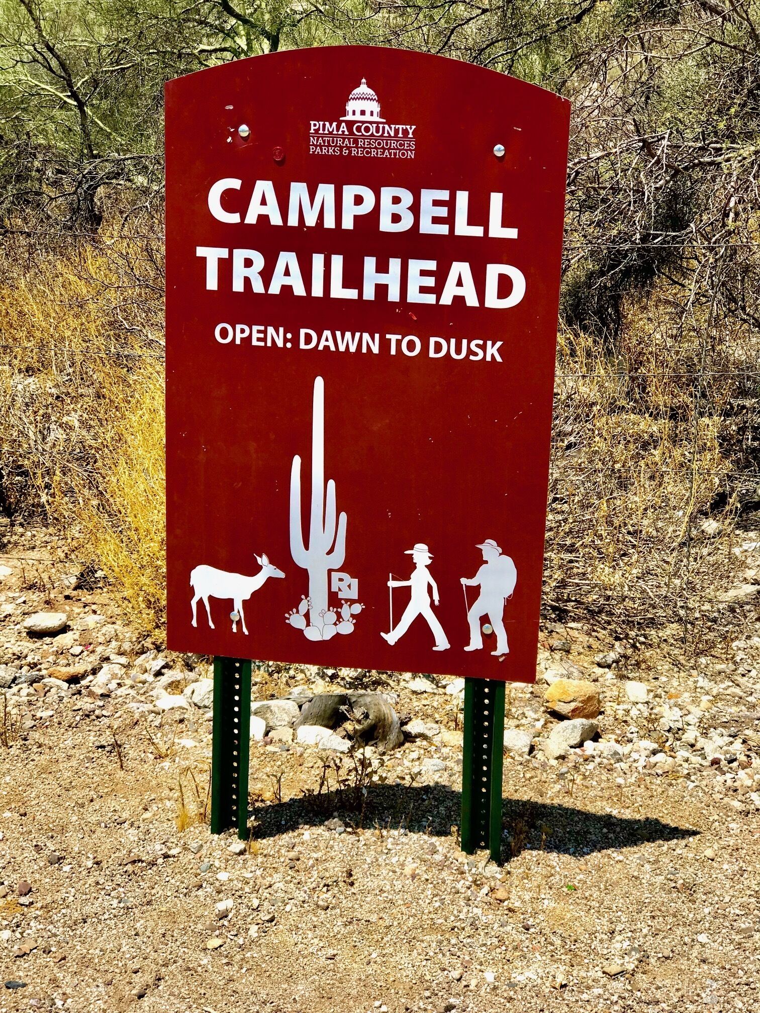 Placa vermelha para Campbell Trailhead, aberta da alvorada ao anoitecer, com símbolos de cavalo, cacto e pessoas caminhando.