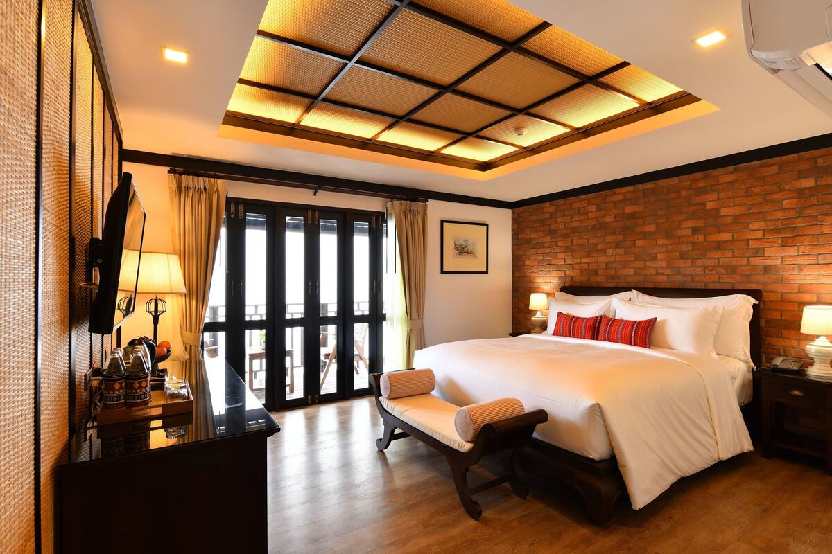 Een hotelkamer met een groot bed, een houten bankje, een bureau met een lamp en uitzicht door een glazen deur.