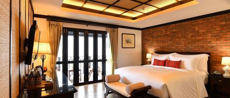 A hotel room with a large bed, a wooden bench, a desk with a lamp, and a view through a glass door.