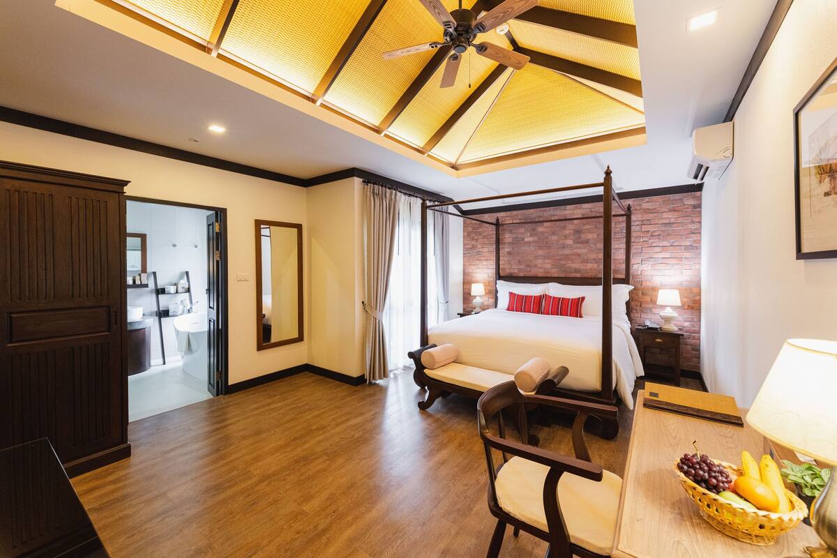 Hotelkamer met een hemelbed, houten vloer, plafondventilator, bureau met een fruitmand, en uitzicht op de badkamer.