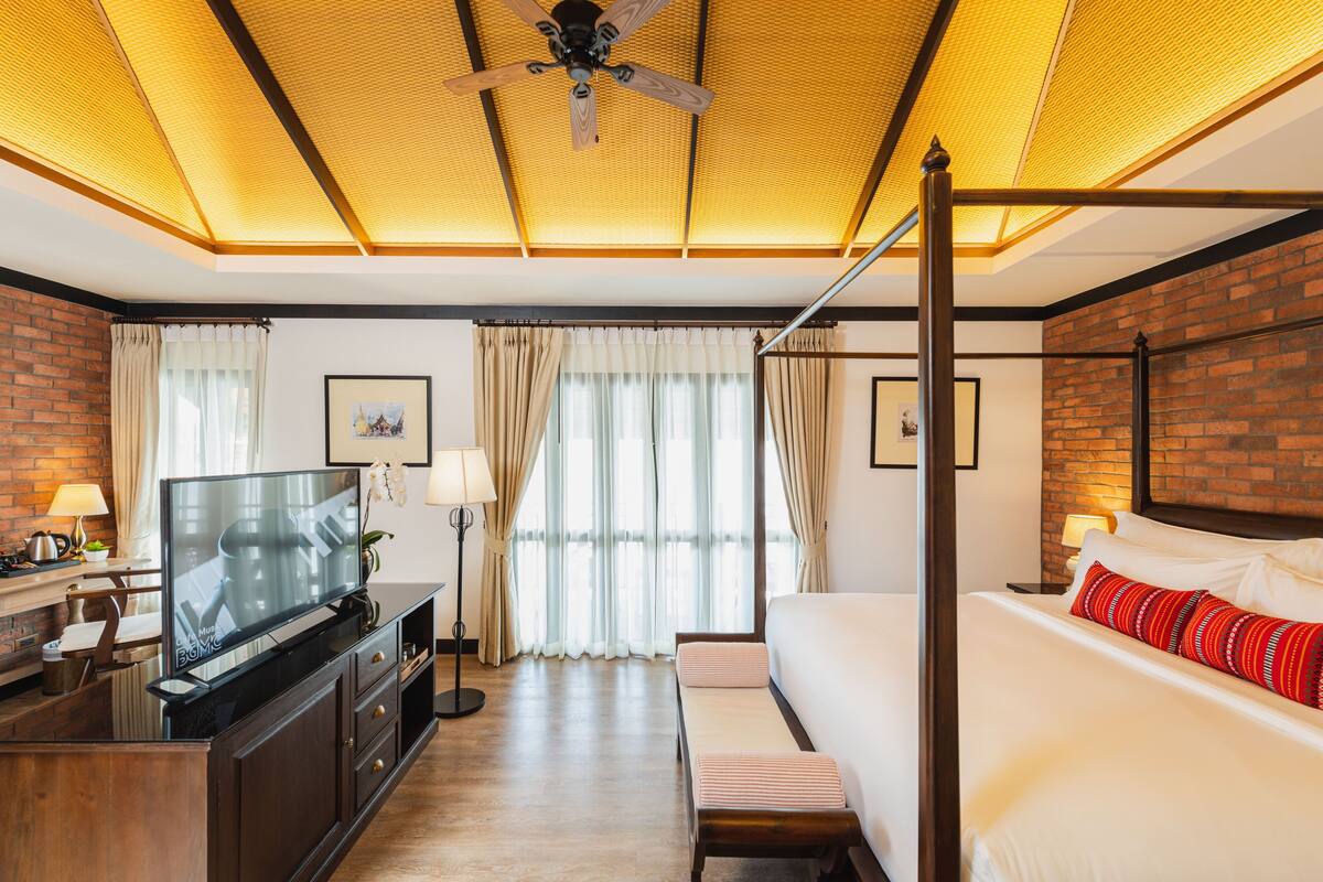 Een hotelkamer met een hemelbed, een flatscreen televisie, een bureau met een lamp en een plafondventilator.