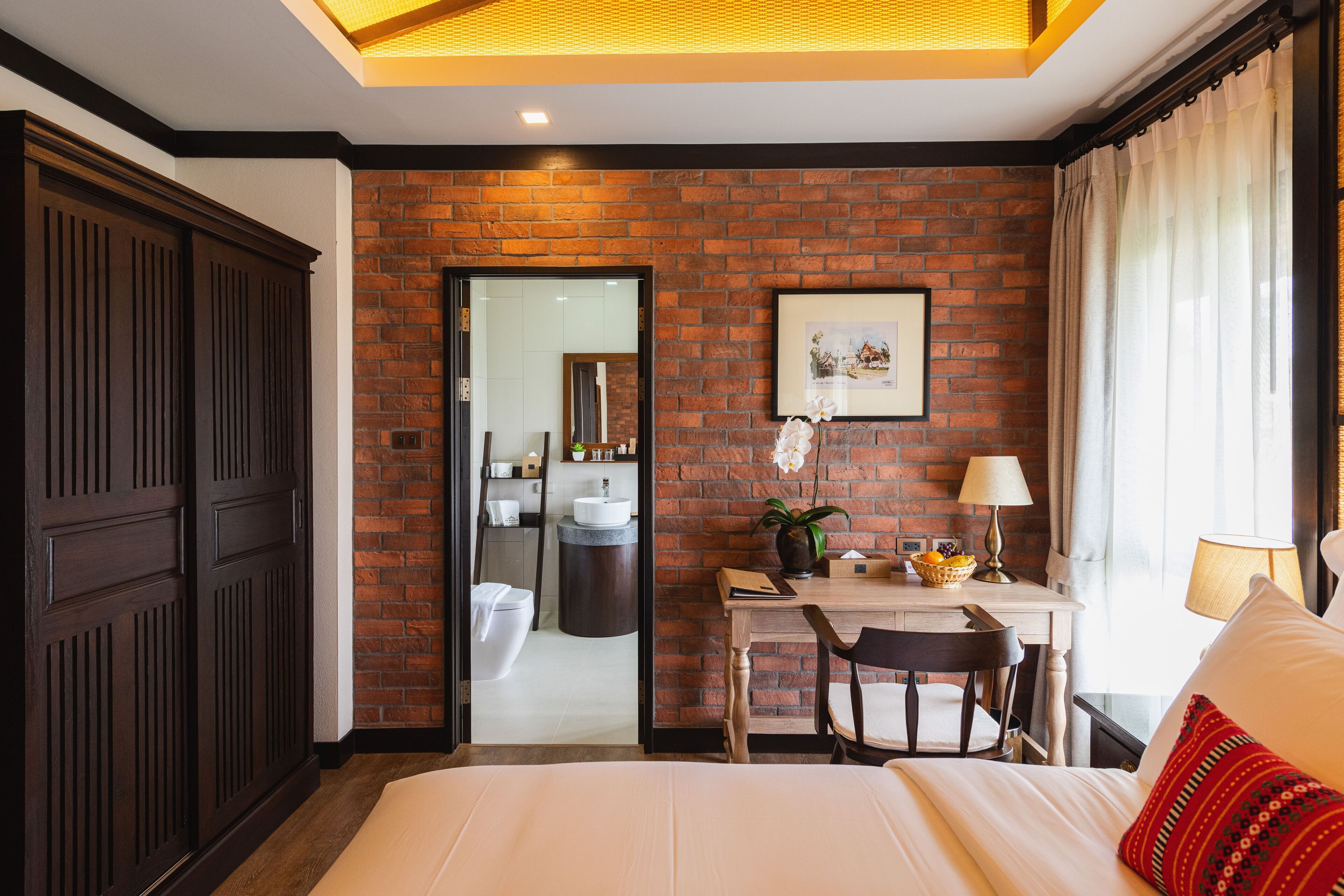 Habitación de hotel con pared de ladrillo, un escritorio de madera, una cama y un baño visible a través de una puerta abierta.