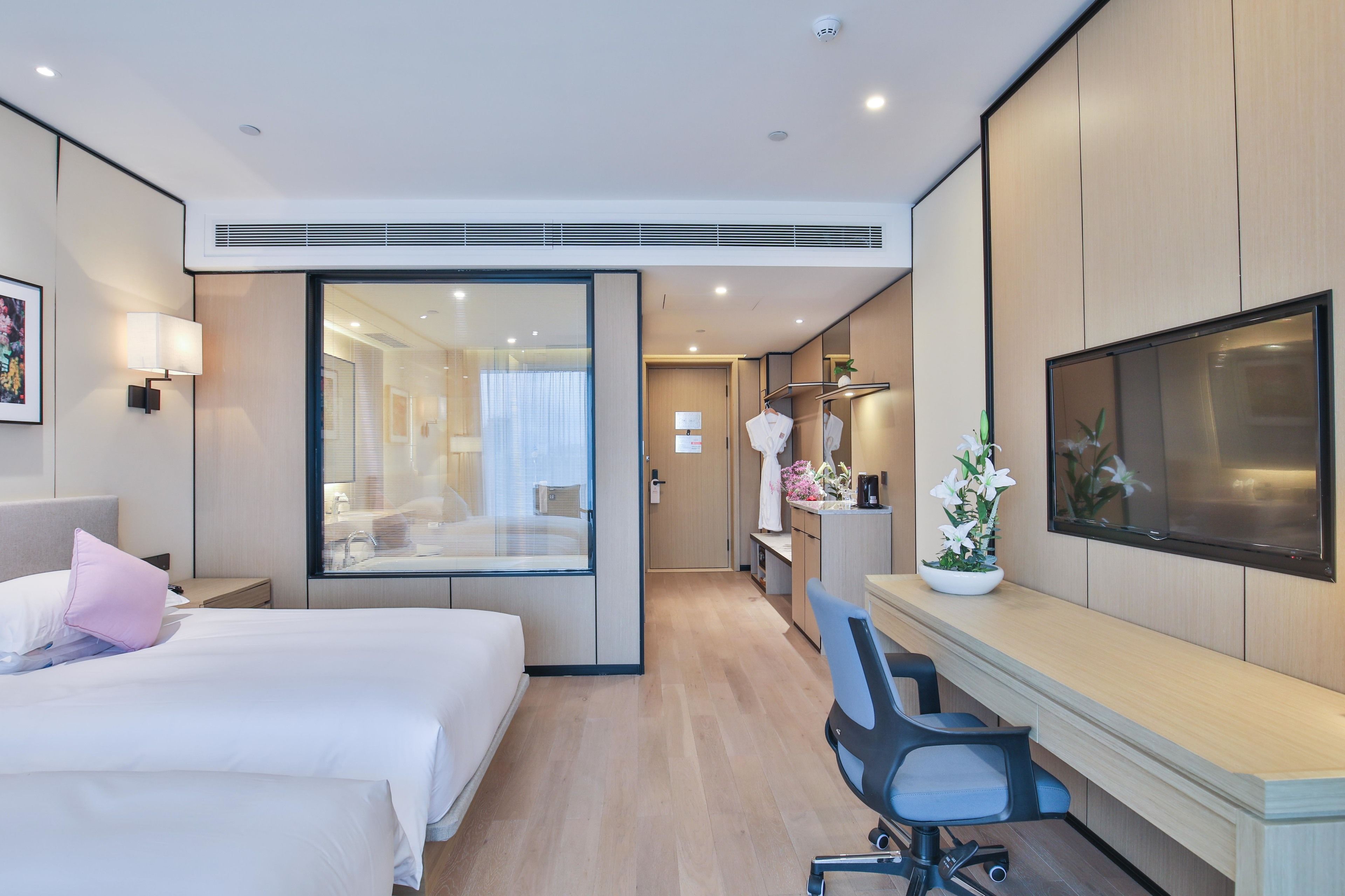 A modern hotel room with a bed, desk, chair, TV, and a bathroom visible through a glass door.