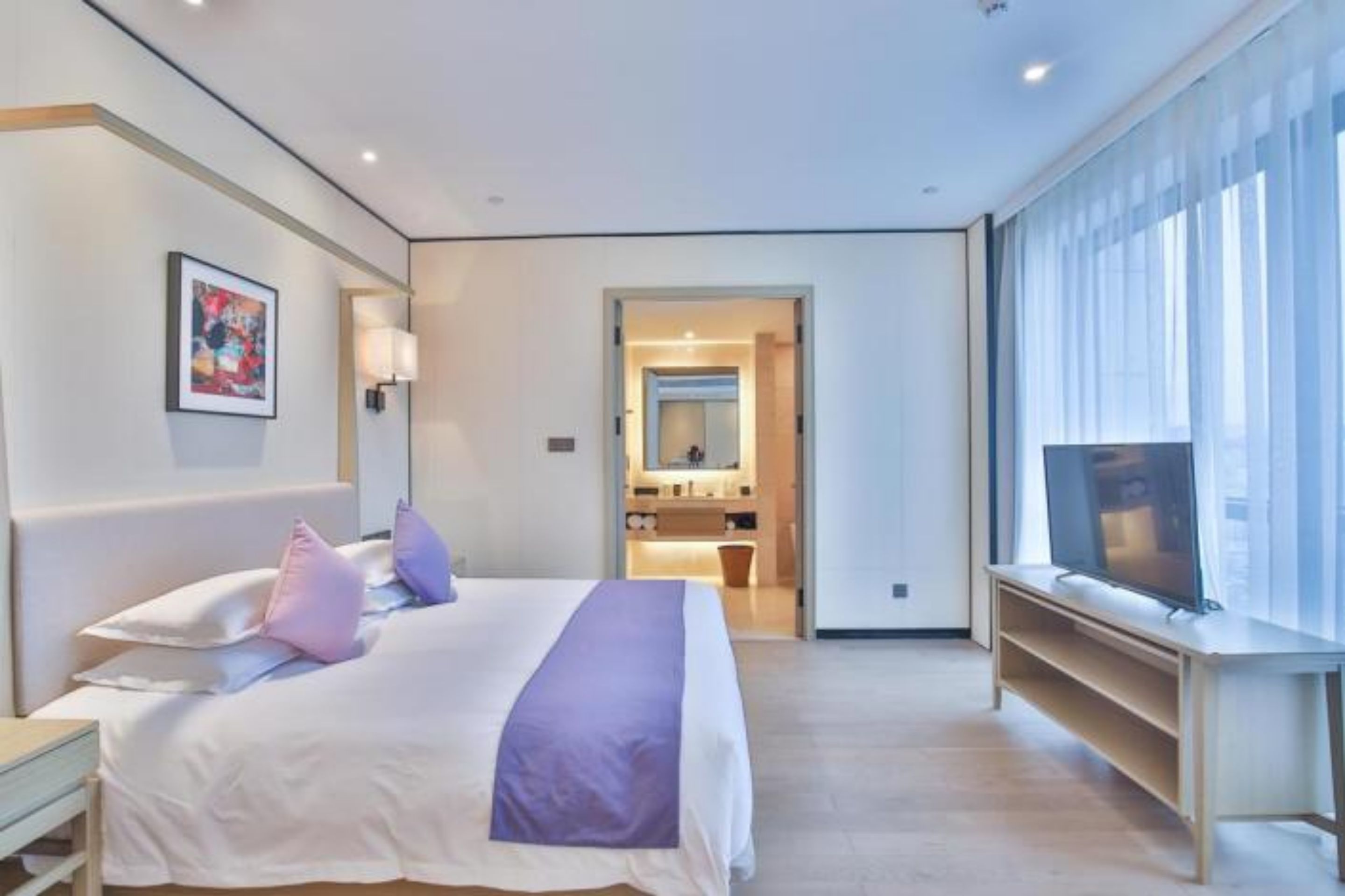 Quarto de hotel moderno com uma cama grande, televisão e banheiro visível por uma porta aberta.