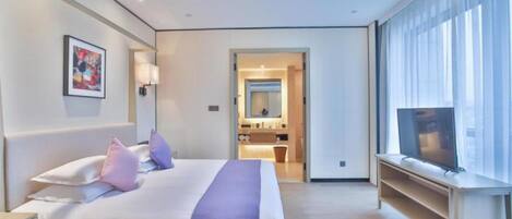 Quarto de hotel moderno com uma cama grande, televisão e banheiro visível por uma porta aberta.