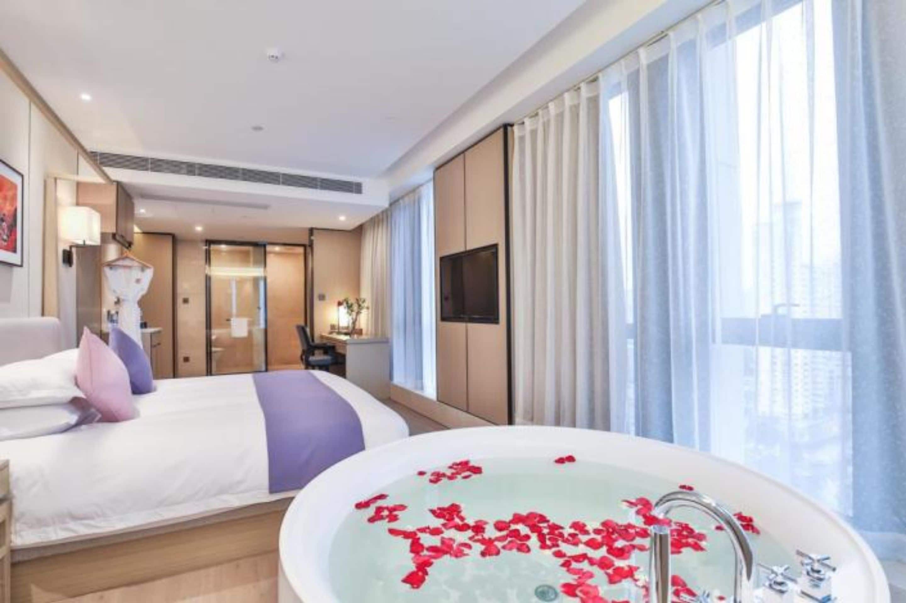 Una habitación de hotel moderna con una cama grande, un área de descanso, un baño con bañera llena de pétalos de rosa y ventanas amplias con cortinas traslúcidas.