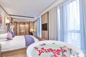 Una habitación de hotel moderna con una cama grande, un área de descanso, un baño con bañera llena de pétalos de rosa y ventanas amplias con cortinas traslúcidas.