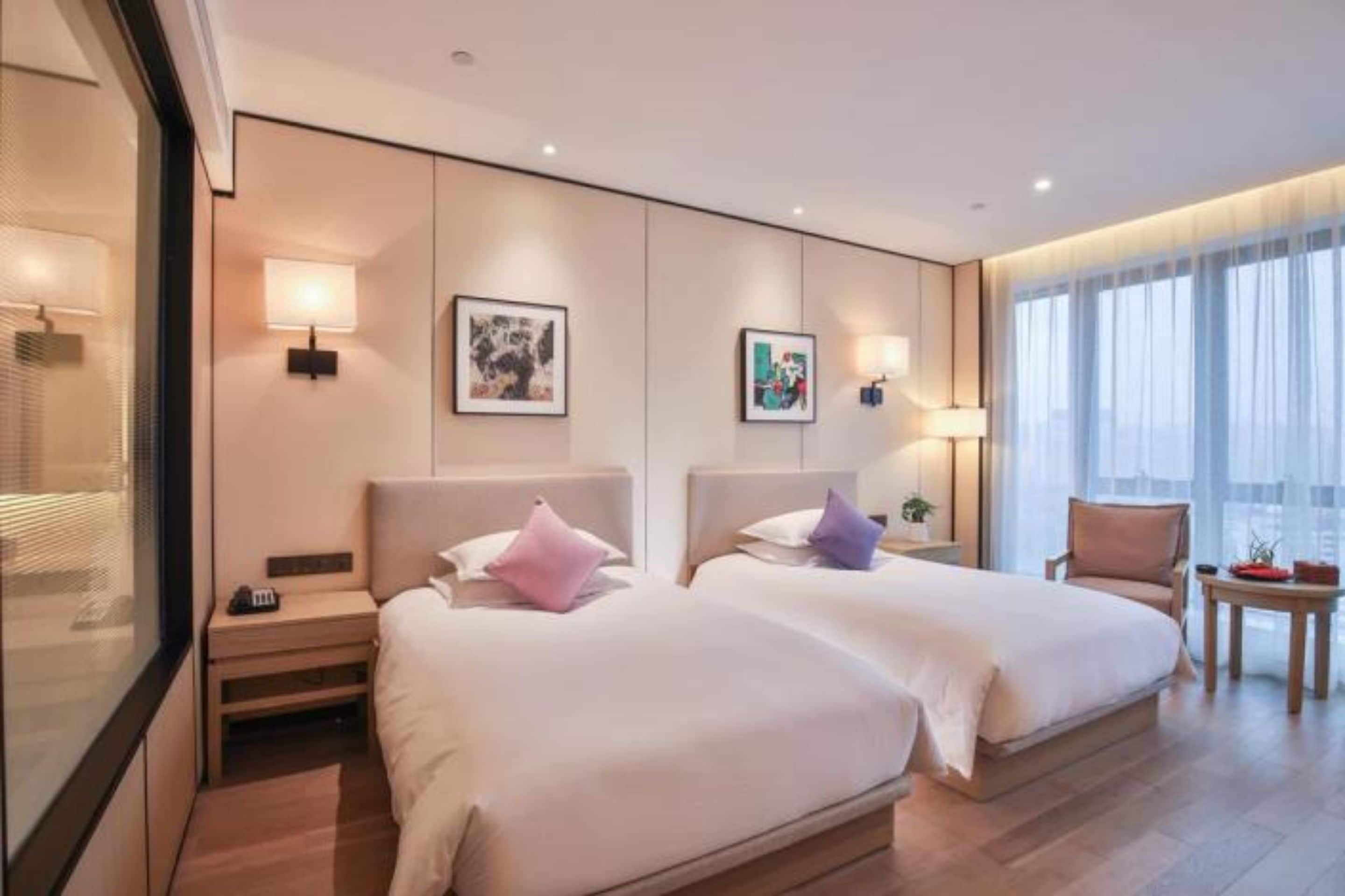 Habitación de hotel con dos camas, mesitas de noche, lámparas de pared, obra de arte enmarcada, una silla y una pequeña mesa con una planta.