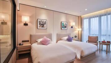 Een hotelkamer met twee bedden, nachtkastjes, wandlampen, een kunstwerk in een lijst, een stoel en een tafeltje met een plant.