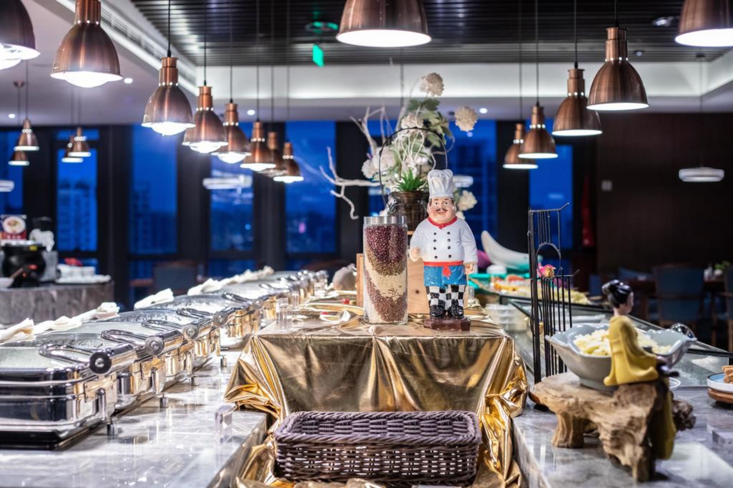 A restaurant setting with a long table covered in a golden cloth, featuring a decorative centerpiece with a chef figurine, a vase with flowers, and a woven basket.