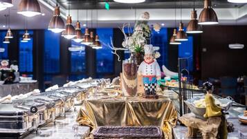A restaurant setting with a long table covered in a golden cloth, featuring a decorative centerpiece with a chef figurine, a vase with flowers, and a woven basket.