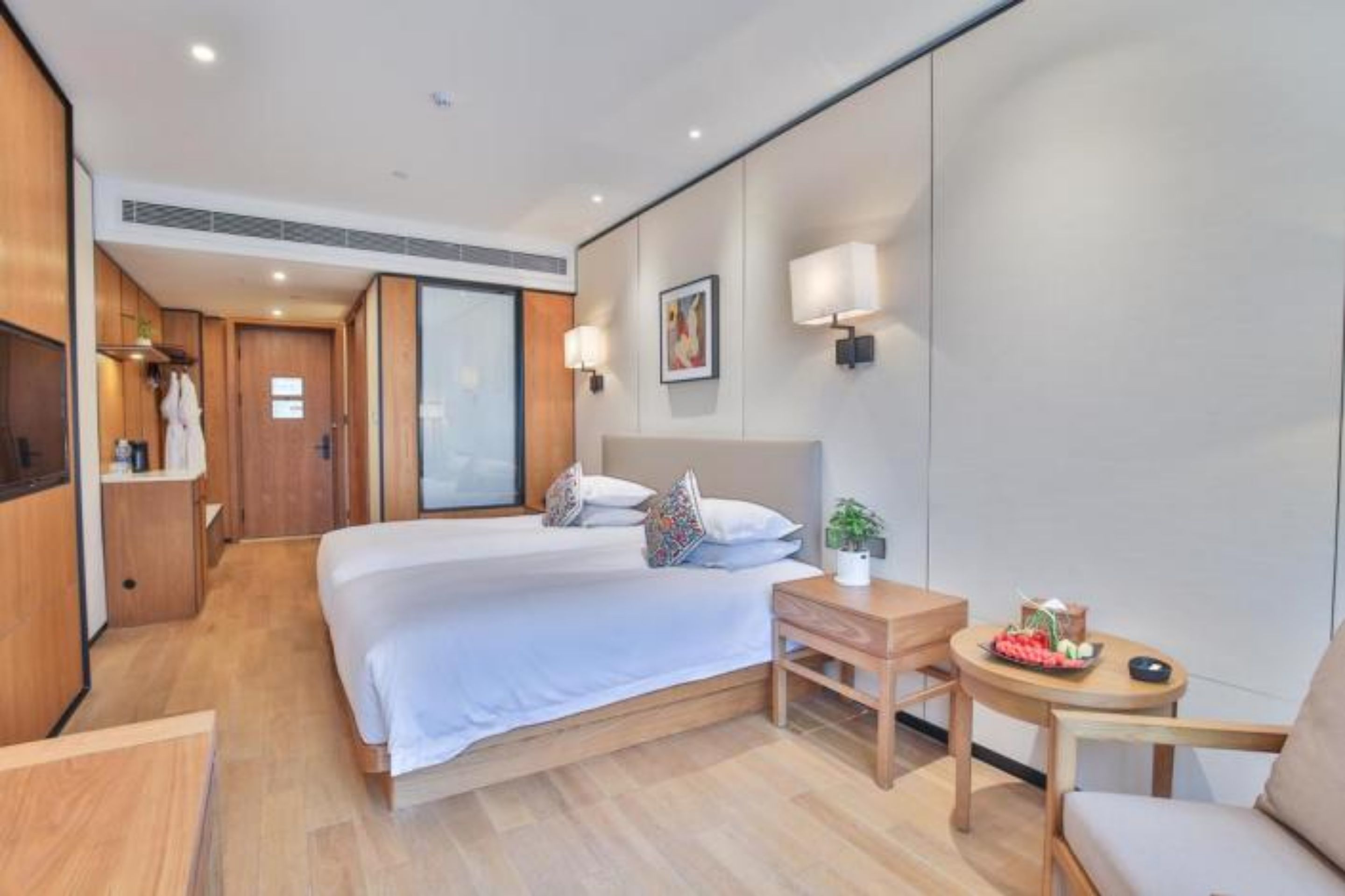 Una habitación de hotel moderna con una cama grande, una mesita de noche con una planta, una pequeña mesa redonda con un frutero y un armario con ropa colgada.