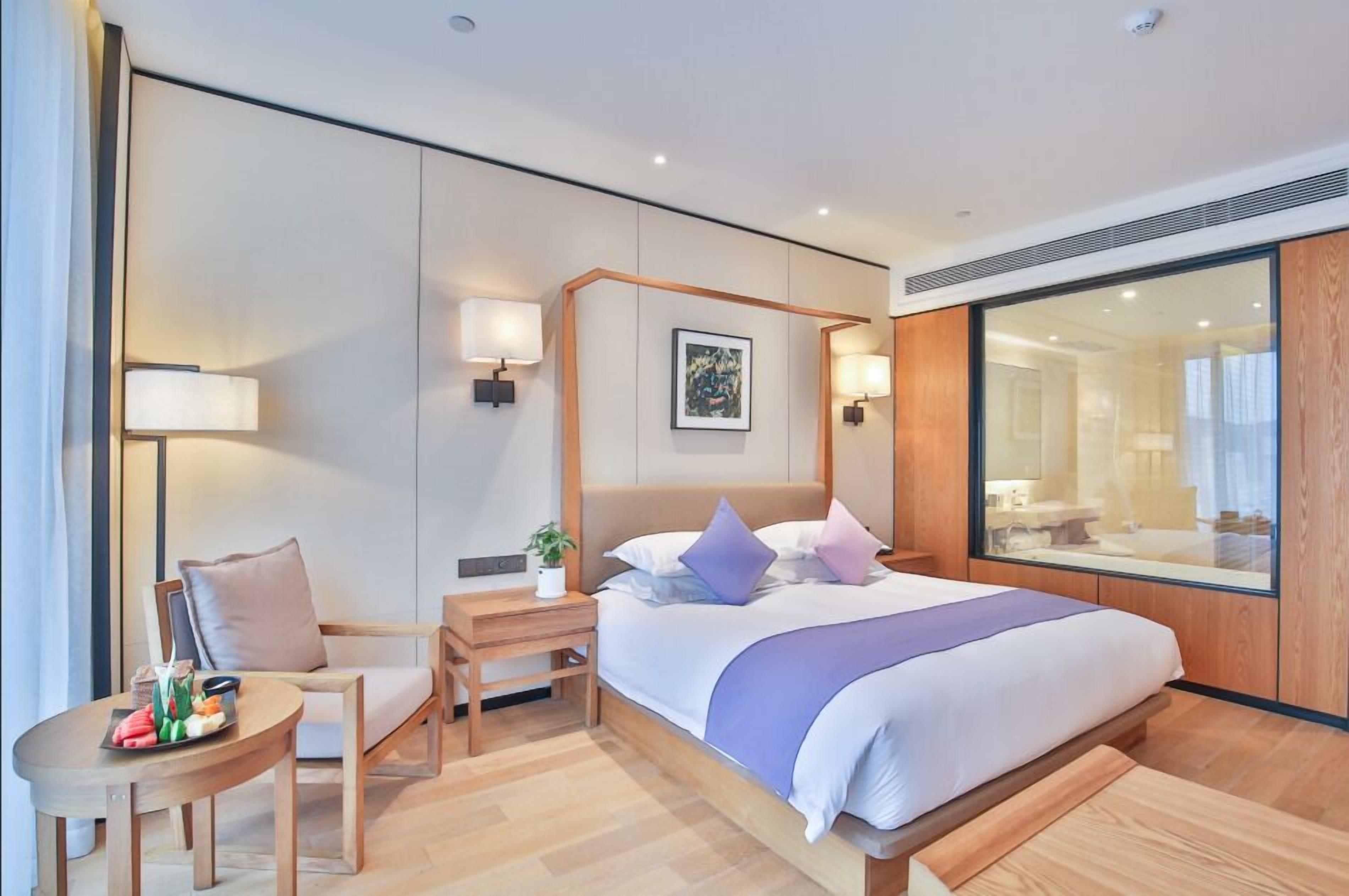 A modern hotel room with a large bed, a sitting area with a chair and table, a bedside lamp, and a large mirror.